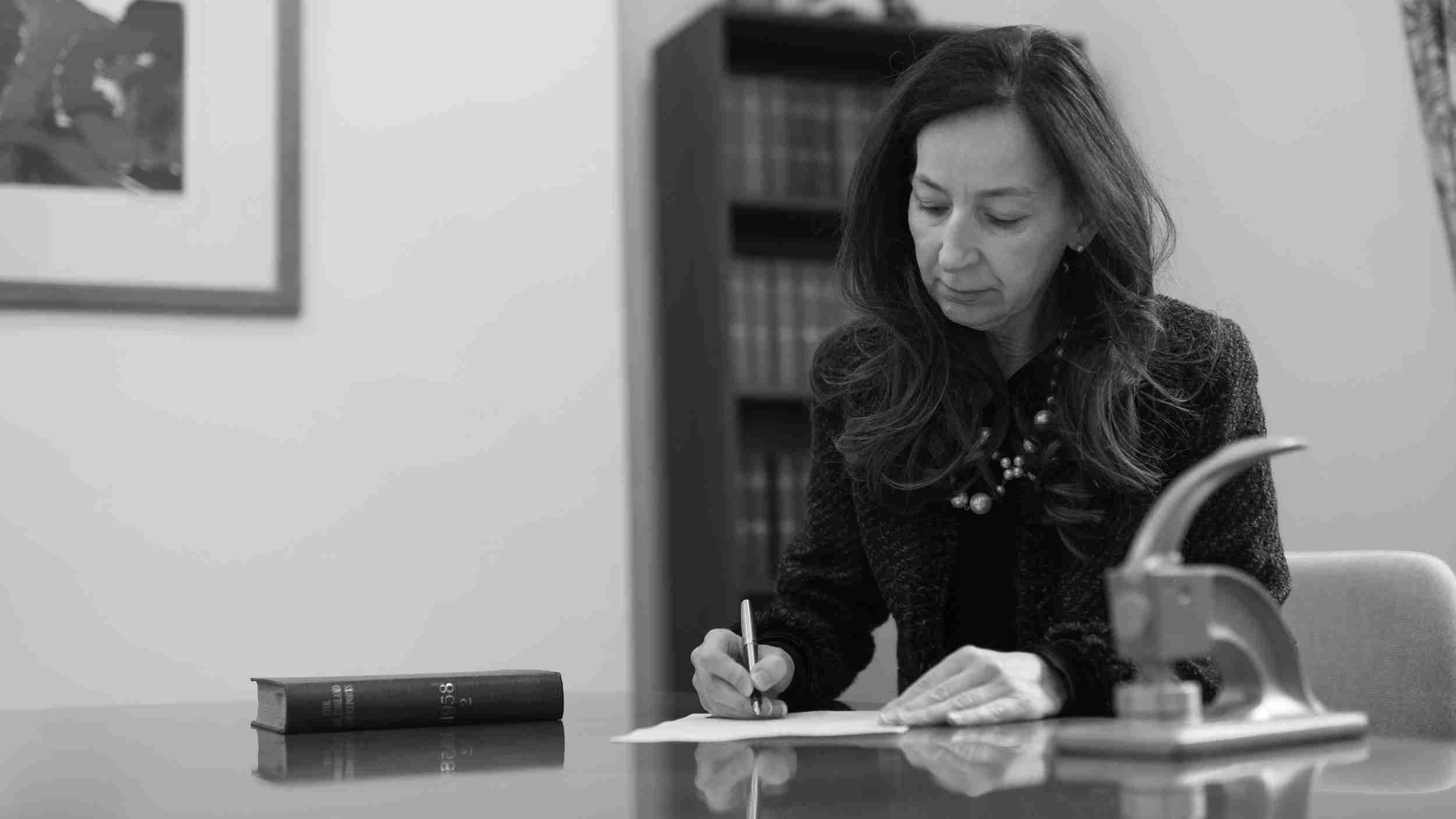 A Davis Gregory director-solicitor at her desk in the Rodney Road office, signing a document beside a 1958-bound legal volume.