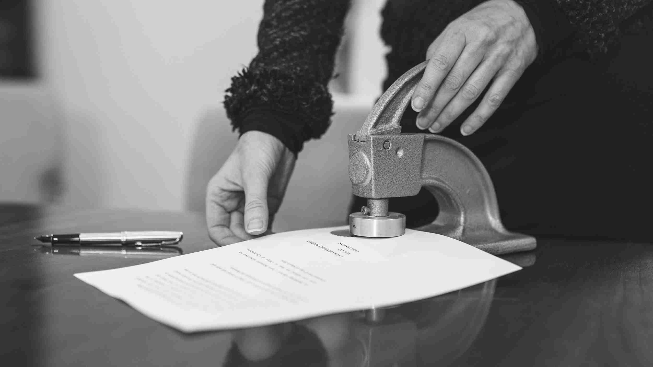 A hand pressing a notarial embosser into a document bearing the Laura Berenice Srodon Cheltenham notary seal, on a polished desk at 25 Rodney Road.
